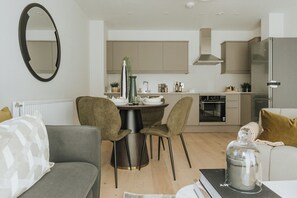 A modern living room with a sofa, a dining table with chairs, a kitchen area with cabinets and appliances, and a large mirror on the wall. - Boutique Luxury Apartment – 2 Bedroom, Sleeps 6 (London)