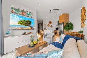 A modern living room with a large flat-screen TV, a wooden coffee table, a beige sofa, and a kitchen area in the background. - Top Floor Beach Condo|Near Eagle Beach|Pool&Jacuzzi (Pos Abou)