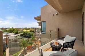 Un balcón con baranda de vidrio, una zona de estar con dos sillas y una mesita, y vistas de los edificios y la vegetación circundantes.
