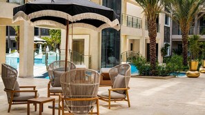 A poolside area with wicker chairs and a large umbrella, surrounded by palm trees and a modern building. - Top Floor Beach Condo|Near Eagle Beach|Pool&Jacuzzi (Pos Abou)