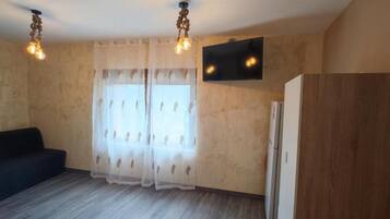 Ein Zimmer mit einer Couch, einem an der Wand befestigten Fernseher, einem Kühlschrank und einem Fenster mit Vorhängen.