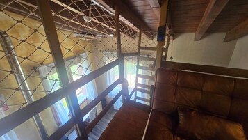 Una habitación con techo de madera, una zona de estar con cojines y vistas a través de una ventana con reja de metal.