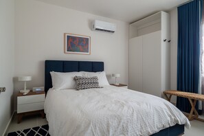 Ein Hotelzimmer mit Bett, Nachttischen, einer fest installierten Klimaanlage, einem gerahmten Kunstwerk und einem Fenster mit Vorhängen.