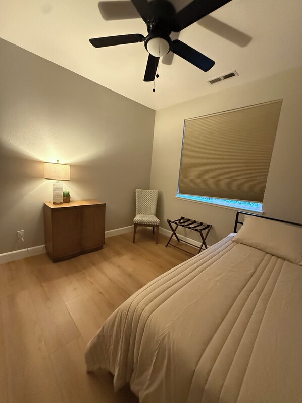 A bedroom with a bed, a ceiling fan, a bedside table with a lamp, a chair, and a window with blinds.