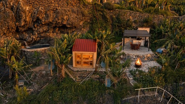 A rustic house with a red roof surrounded by lush vegetation and a stone wall.