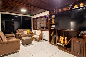 A living room with a sofa set, a chair, a television, and decorative items on shelves. - Casa 1 Porto Seguro 6 Quartos e Piscina (Porto Seguro)