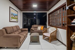 A living room with a sofa, armchair, ottoman, and wicker chair. There is a painting on the wall and a bookshelf with books. - Casa 1 Porto Seguro 6 Quartos e Piscina (Porto Seguro)