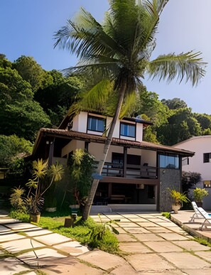 A two-story building with a balcony, surrounded by lush greenery and a palm tree. - Casa 1 Porto Seguro 6 Quartos e Piscina (Porto Seguro)