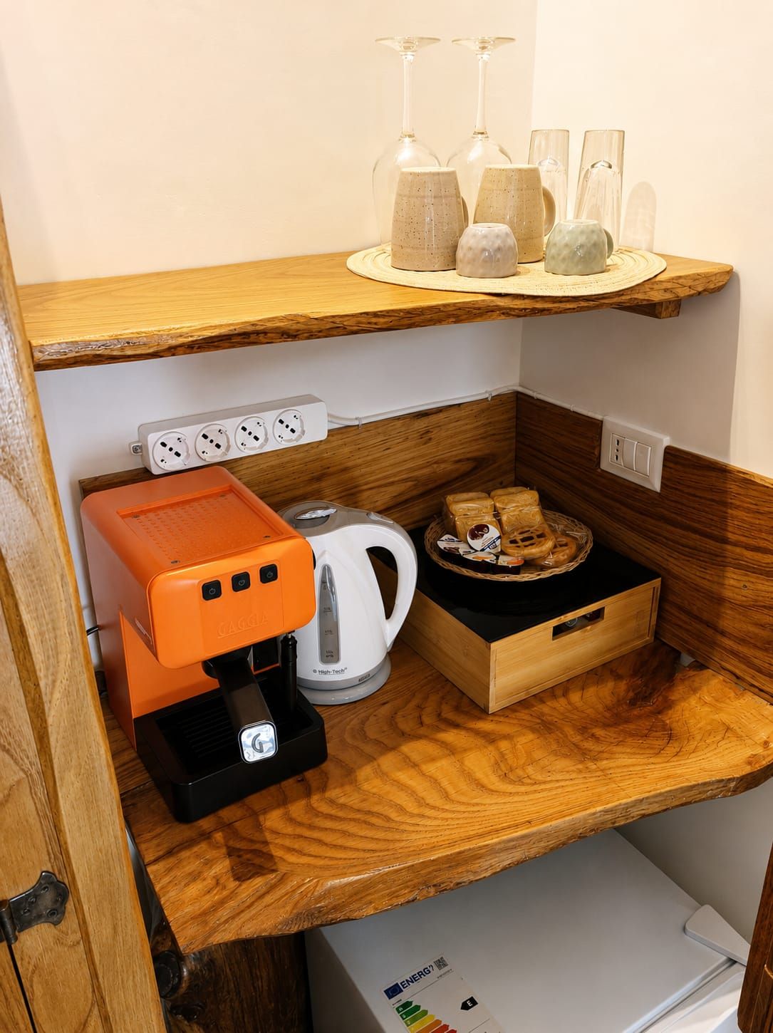 Eine Küchenzeile mit Kaffeemaschine, Wasserkocher und Brötchenkorb auf einer Holzausziehtafel.