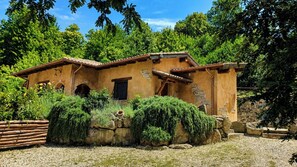 A rustic building with a tiled roof, stone walls, and a garden area with various plants and a wooden fence.