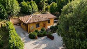 Una casa a un piano con tetto in tegole, circondata da un vialetto sterrato e rigogliosa vegetazione.