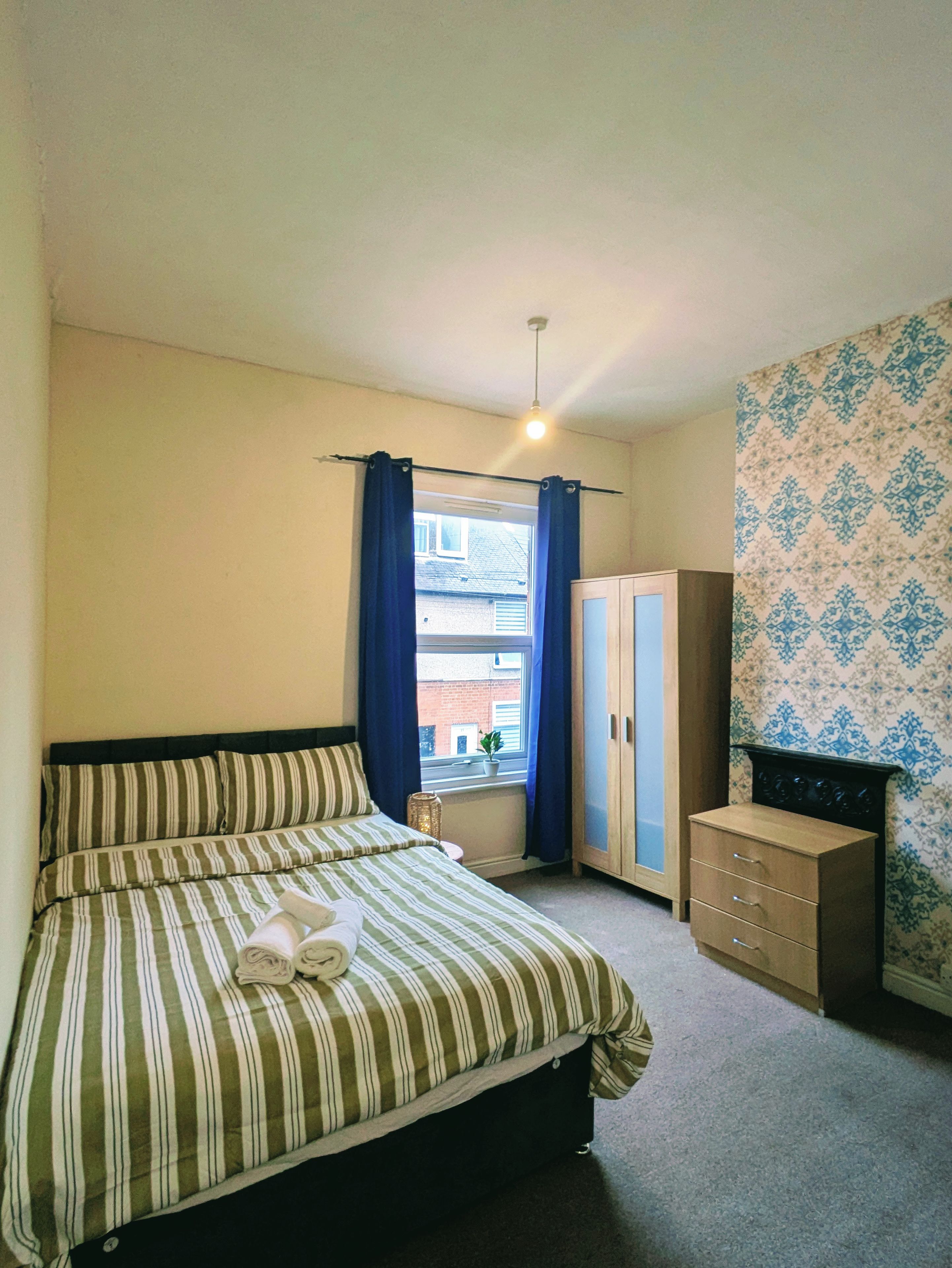 A bedroom with a bed, a wardrobe, a window with curtains, and a patterned wall.