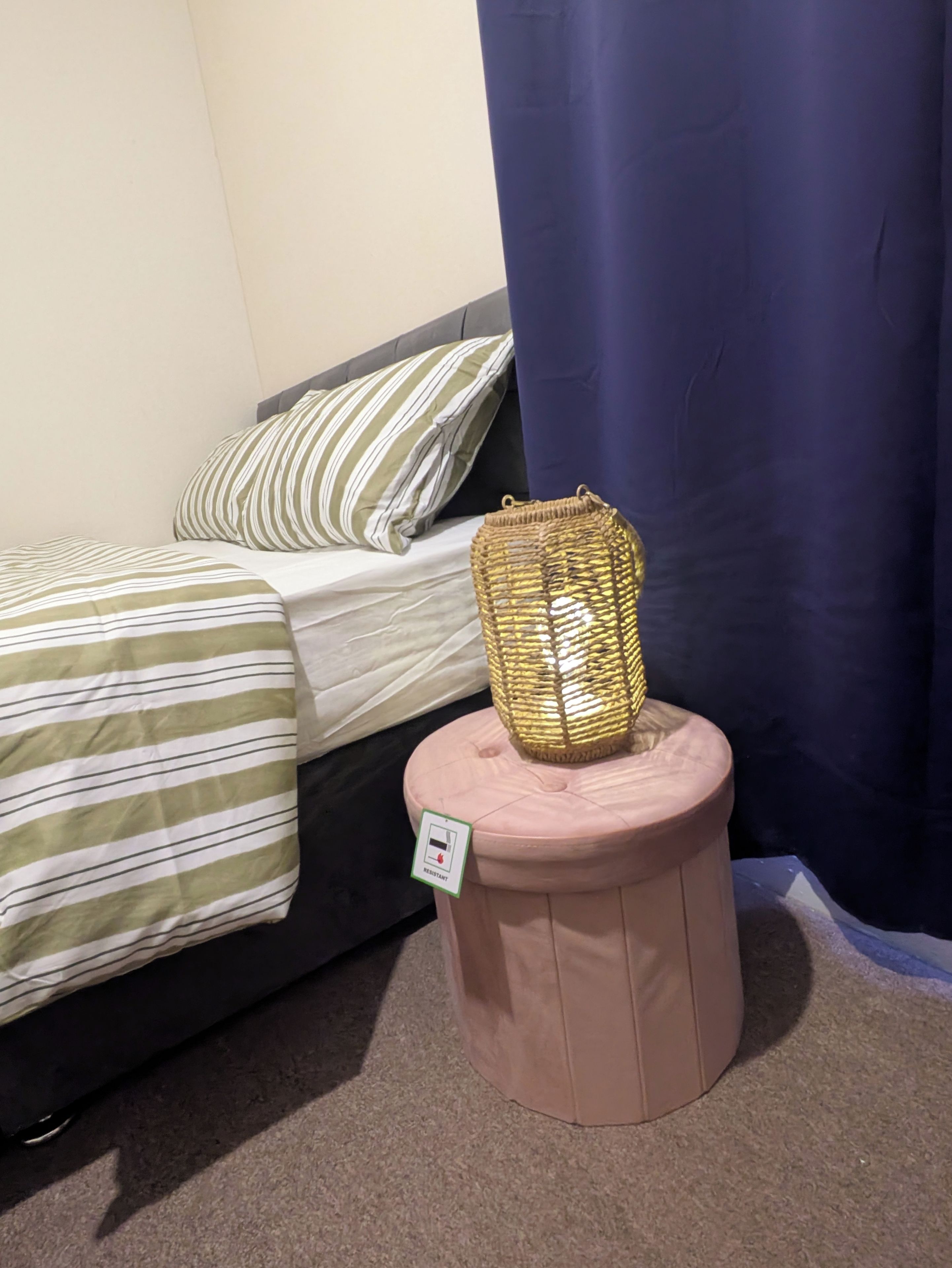 A bedroom with a bed, a striped pillow, a woven basket on a stool, and a blue curtain.