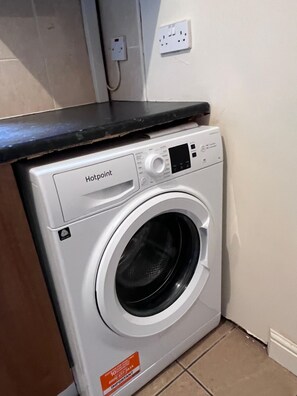 Lavadora Hotpoint de carga frontal con panel de control, ubicada en una esquina de una habitación con piso de baldosas.