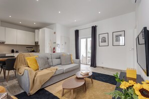 A modern living room with a grey sofa, a coffee table, and a dining area with a table and chairs. There are two framed pictures on the wall, a fire extinguisher, and a window with curtains. - Baixa Garden: 2BR + Patio | Perfect for Groups and Families (Lisboa)