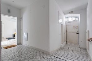 A hallway with patterned floor tiles, a door with a decorative metal grille, and a wall-mounted light switch. - Baixa Garden: 2BR + Patio | Perfect for Groups and Families (Lisboa)