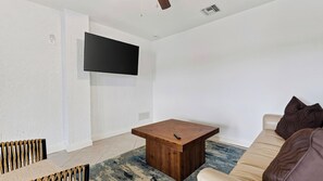 A modern living room with a flat-screen TV mounted on the wall, a wooden coffee table, a beige sofa with brown cushions, and a patterned rug. - Sanctuary Point by Avantstay Waterfront Pool + Spa Kayak + Fishing Dock Near Beaches (Bonita Springs)