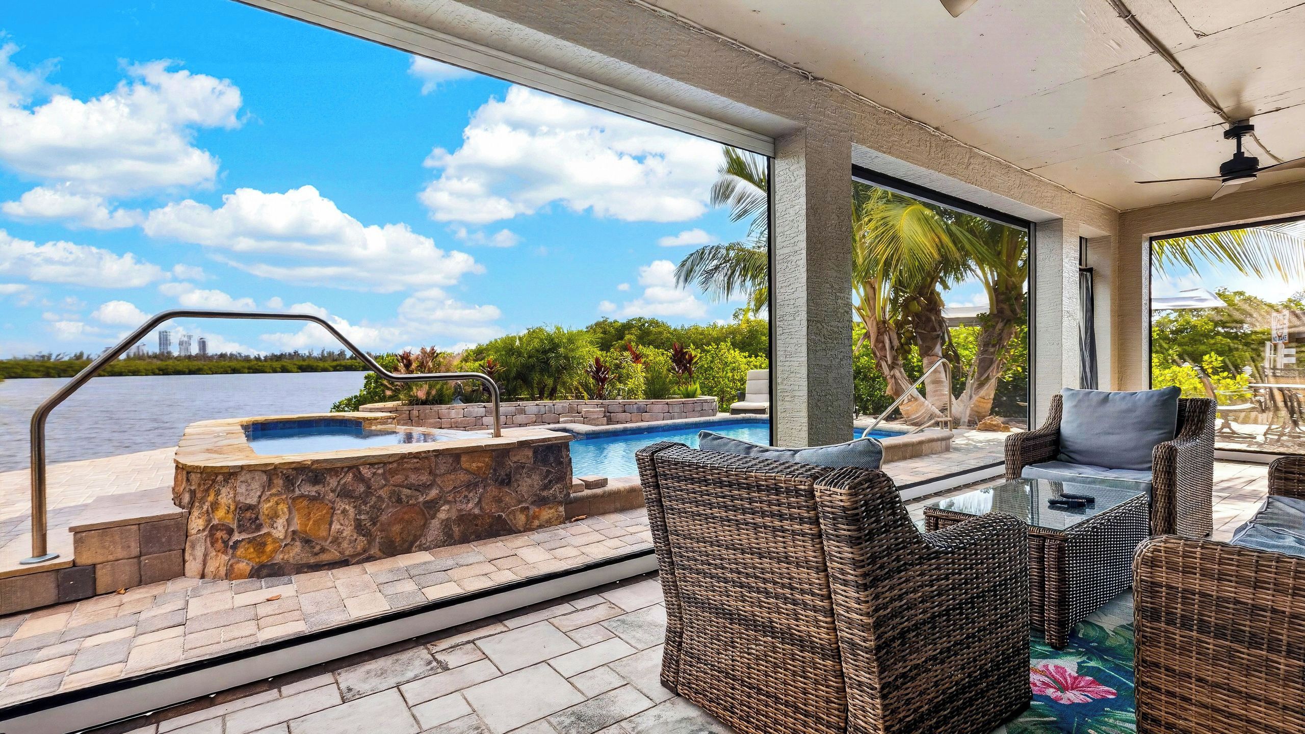 A pool area with a hot tub, wicker furniture, and a view of a body of water and palm trees.