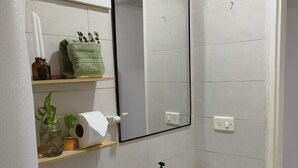 Un baño con lavamanos, inodoro y espejo blancos. Hay repisas con artículos de tocador y una planta en maceta.