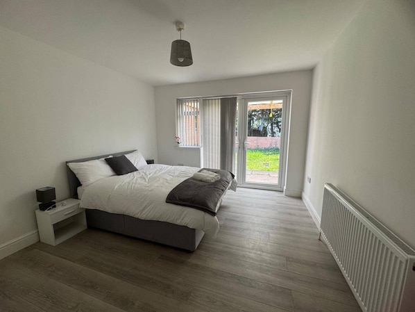 A bedroom with a bed, bedside table, and a view of the outdoors through a sliding door. - Apartment Sleeps 7 With Patio (Hayes)