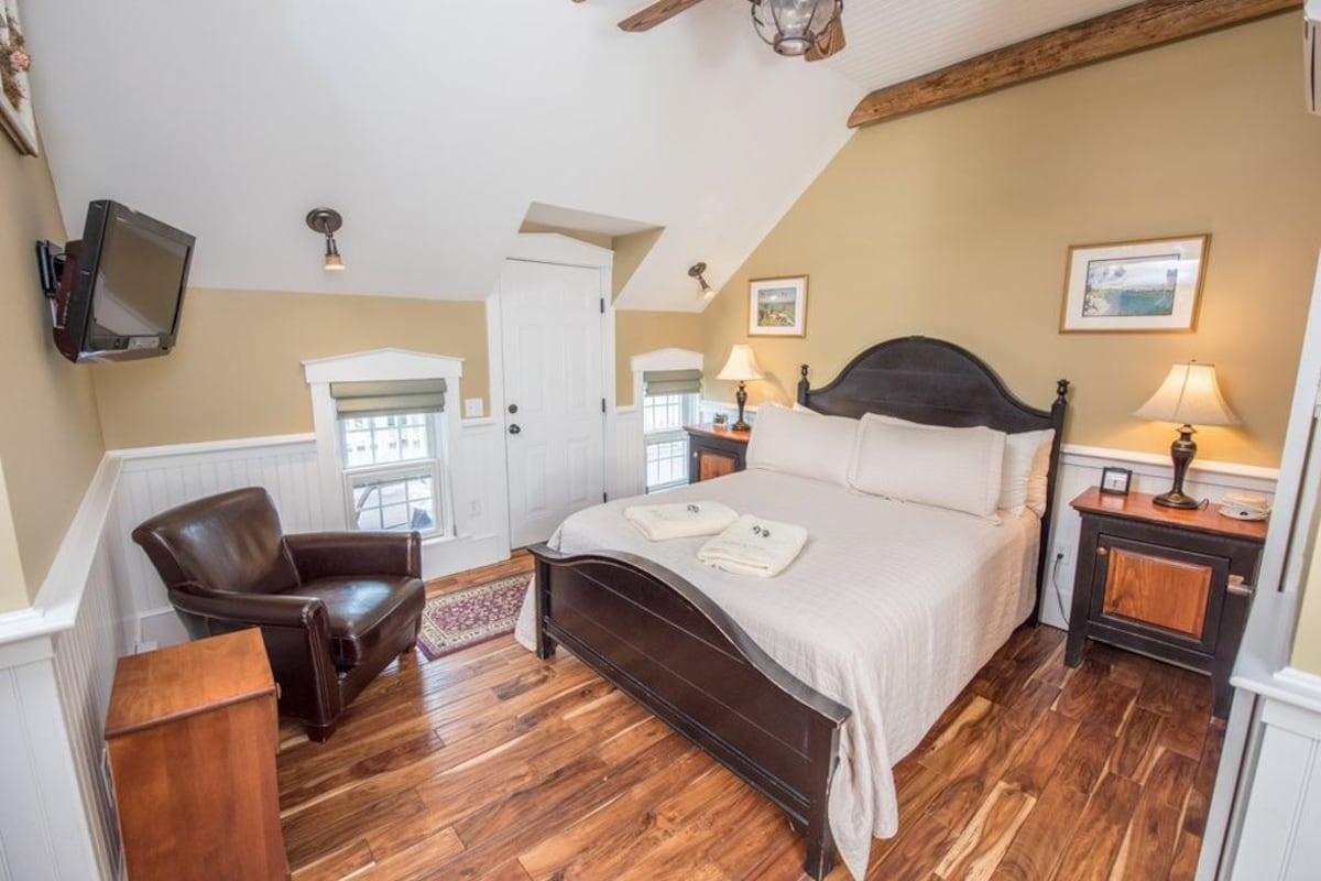 Quarto com piso de madeira, cama com cabeceira escura, uma cadeira, mesa de cabeceira e televisão na parede.
