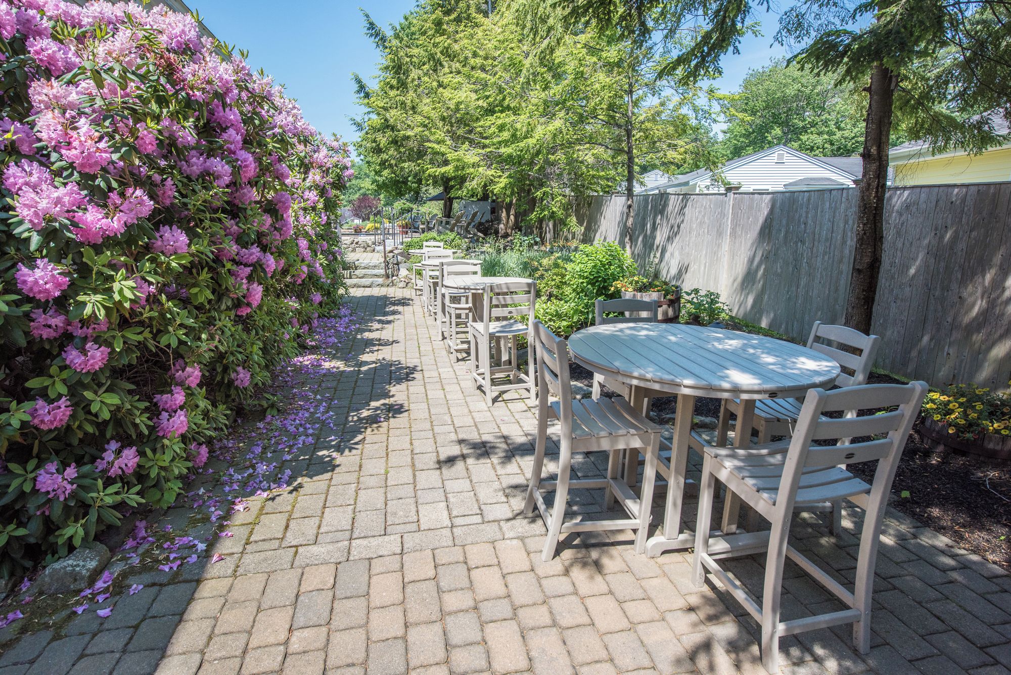 Un patio con una mesa redonda y sillas, rodeado por un camino de ladrillo y arbustos en flor.