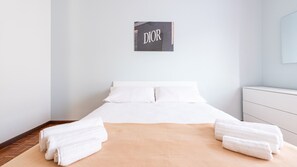 Een hotelkamer met een bed, witte lakens, een beige plaid en een bord aan de muur met de tekst 'DIOR'.