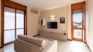 Een woonkamer met een bank, een flatscreen televisie, een wandplank en een raam met uitzicht op een stadsbeeld.