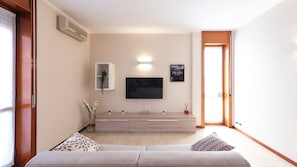 Una sala de estar con un sofá, un televisor de pantalla plana montado en la pared, un mueble bajo con una planta y una puerta que da a otra habitación.