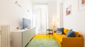 Ein modernes Wohnzimmer mit einem Fernseher auf einem weißen Schrank, einem gelben Sofa mit blauen Kissen, einem kleinen Beistelltisch und einer Stehlampe.