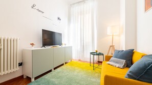 A modern living room with a flat-screen TV on a cabinet, a yellow sofa with blue cushions, a small side table, and a floor lamp. - [Corso Buenos Aires] Central and Cozy (Milano)