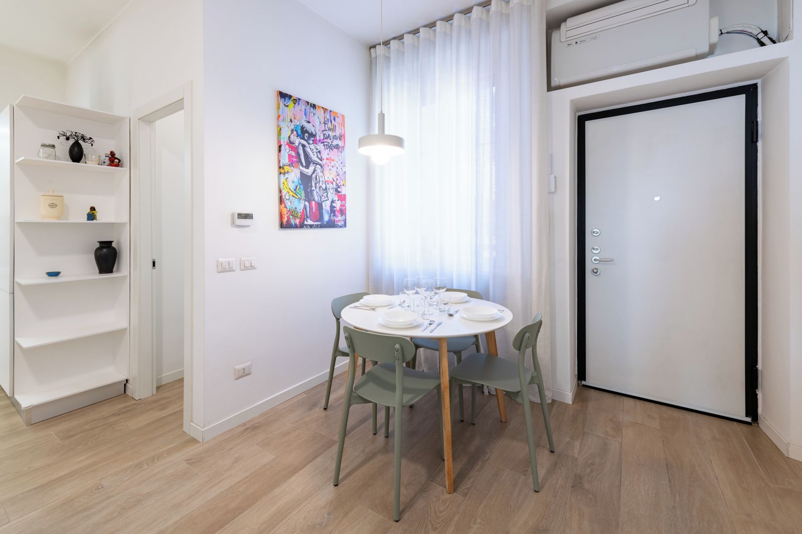 A dining area with a round table set for four, a white door, and a shelf with decorative items.