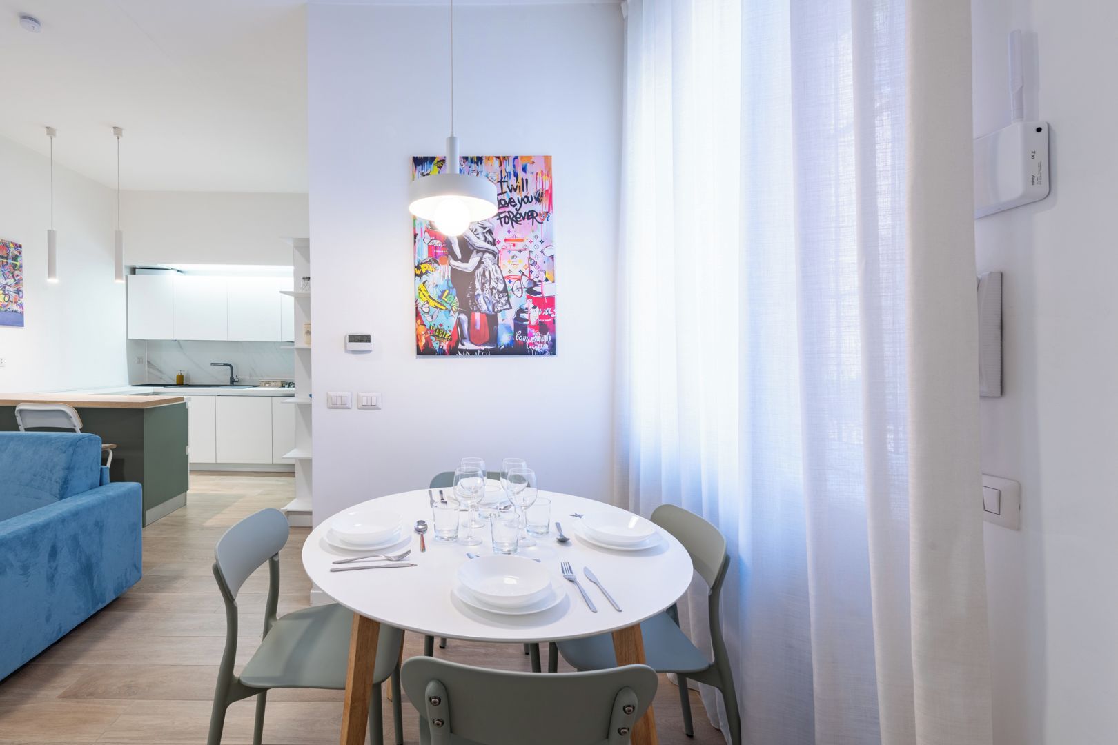 Ein moderner Essbereich mit einem runden Tisch, gedeckt für vier Personen, einem blauen Sofa und einem farbenfrohen, abstrakten Gemälde an der Wand.