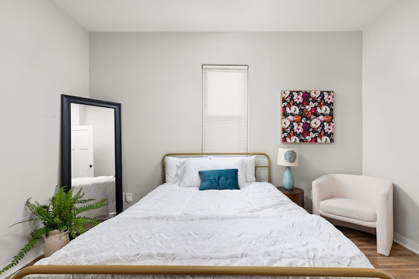 A bedroom with a bed, a chair, a mirror, a window with blinds, and a floral artwork on the wall. - 2-bedroom house in peaceful Indianapolis with cozy vibes (Indianapolis)
