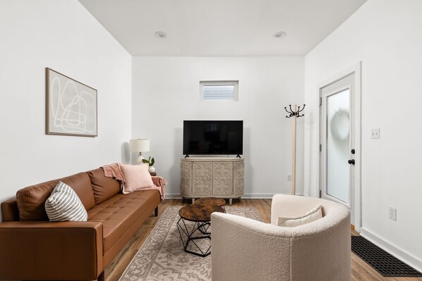 Een moderne woonkamer met een bruine leren bank, een witte fauteuil, een televisie op een kast en een kapstok.