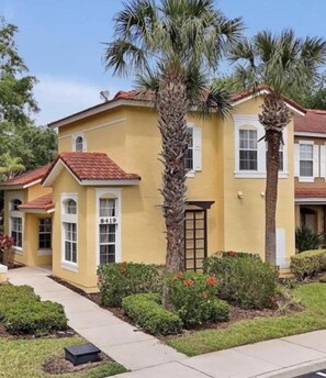 A yellow two-story house with a red roof, a number '8419' on the front, and a palm tree in the front yard. - 4BR Townhouse- Near Disney (Kissimmee)