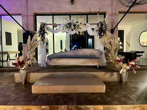 A bed with a mattress on a platform, surrounded by decorative plants and a white draped fabric.