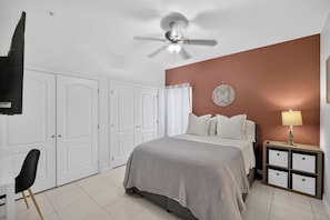 Un dormitorio con una cama, una mesita de noche, una lámpara, un ventilador de techo y puertas blancas.