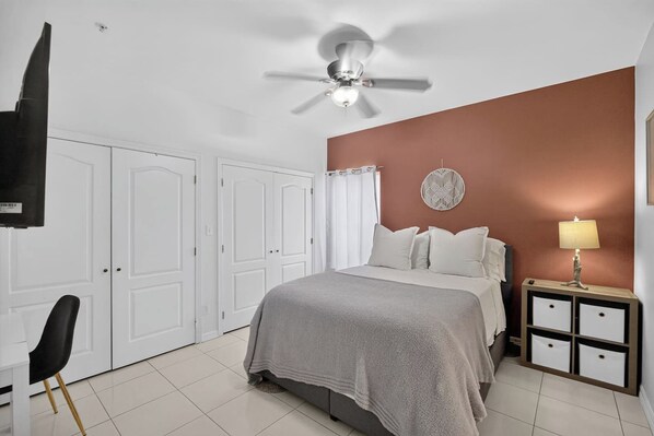 Quarto com cama, mesa de cabeceira, abajur, ventoinha no teto e portas brancas.