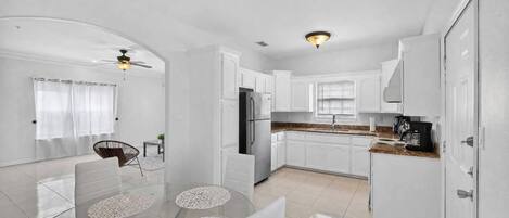 Una cocina moderna con armarios blancos, un comedor con mesa y sillas de vidrio y un ventilador de techo.