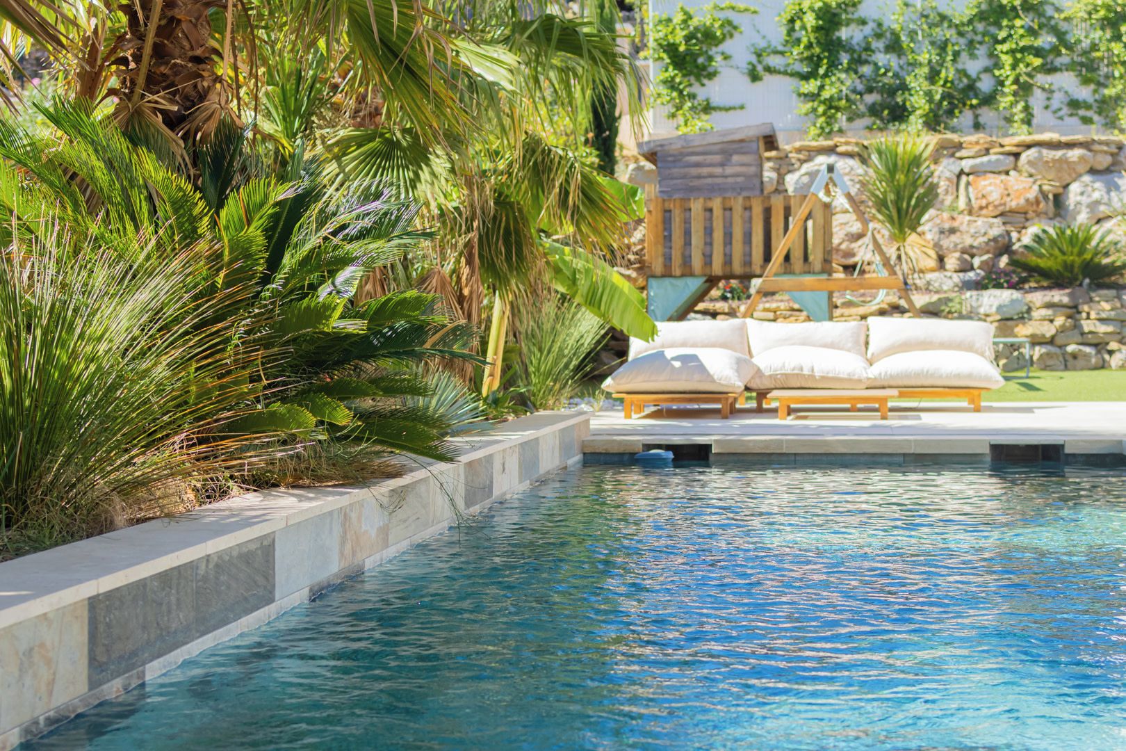 A swimming pool with a wooden deck and lounge chairs, surrounded by tropical plants and a stone wall.