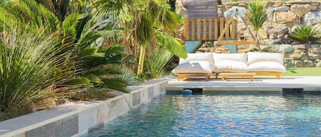 A swimming pool with a wooden deck and lounge chairs, surrounded by tropical plants and a stone wall.