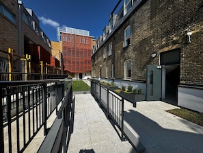 A corridor with a modern building featuring a red lattice facade, a brick building with a door, and a clear sky. - Stylish 1-bed Apt in Clapham - 1 Minute from tube (London)