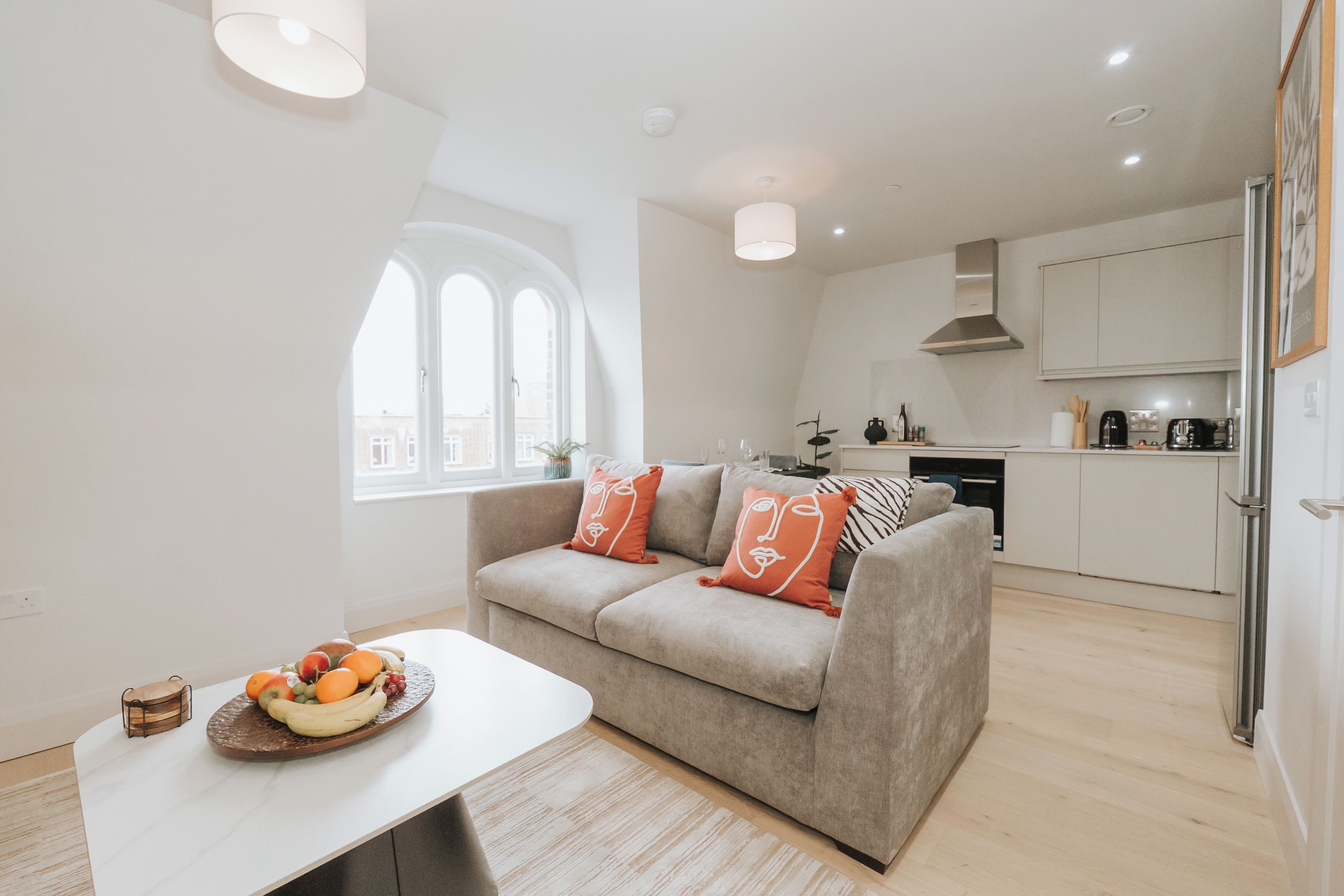 A modern living room with a sofa, a dining table with fruit, and a kitchen in the background.