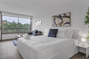 A modern bedroom with a large bed, a seating area, and a view through a large window. - Cozy Nook Studio 8 min to Disney & Universal | Orl (Kissimmee)