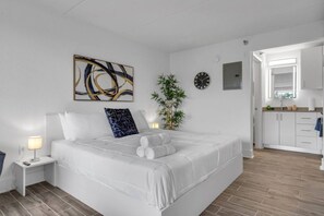 Ein Hotelzimmer mit einem Bett, einem Nachttisch mit Lampe, einer Wanduhr, einer Topfpflanze und Blick in die angrenzende Küche.