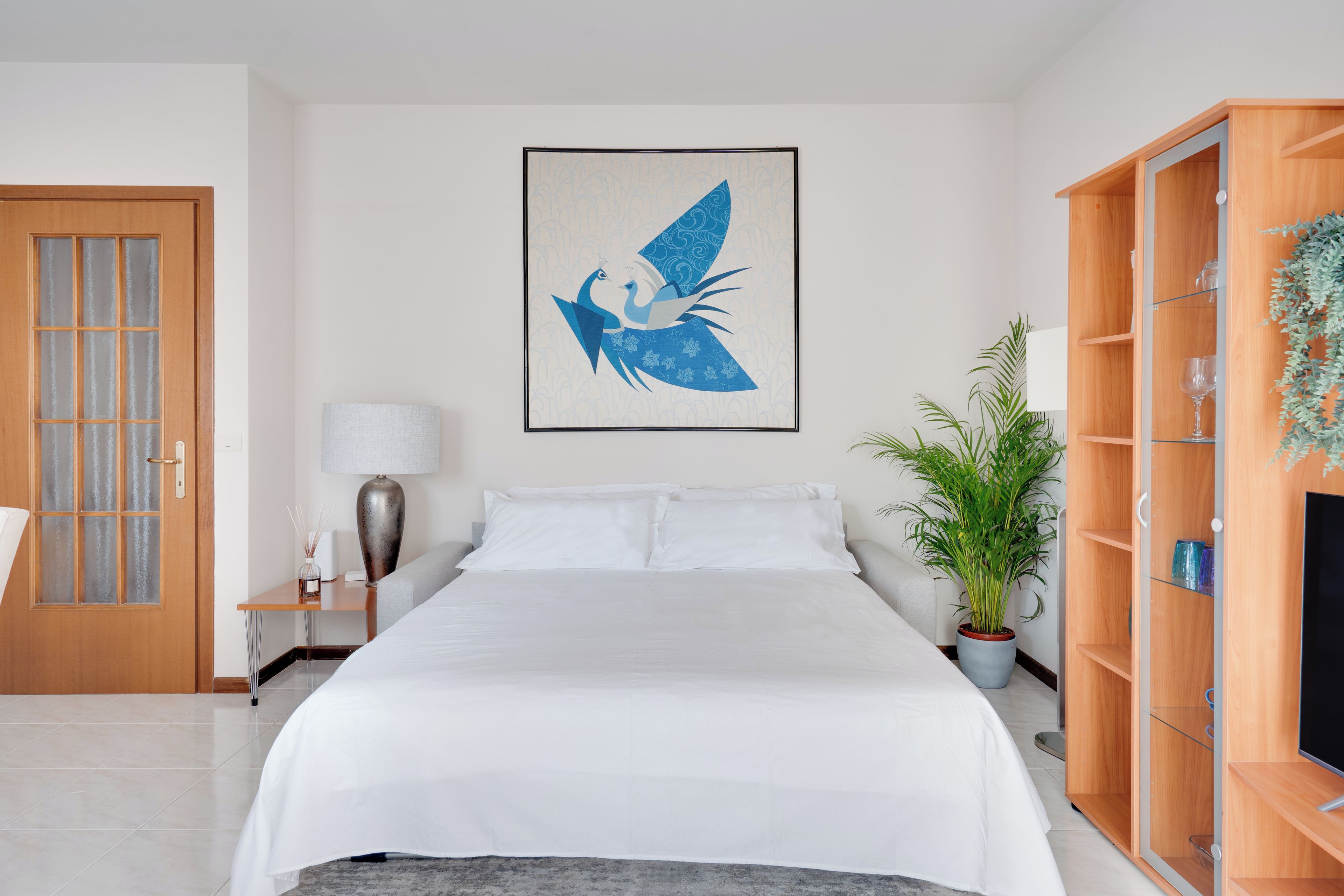 A hotel room with a bed, a nightstand, a lamp, a potted plant, and a wooden cabinet with shelves.