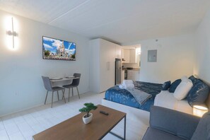 A modern, compact living space with a dining area, a bed with a blue floral bedspread, and a small potted plant on a coffee table. - Breezy Studio 8 min to Disney + Parking | Orl (Kissimmee)