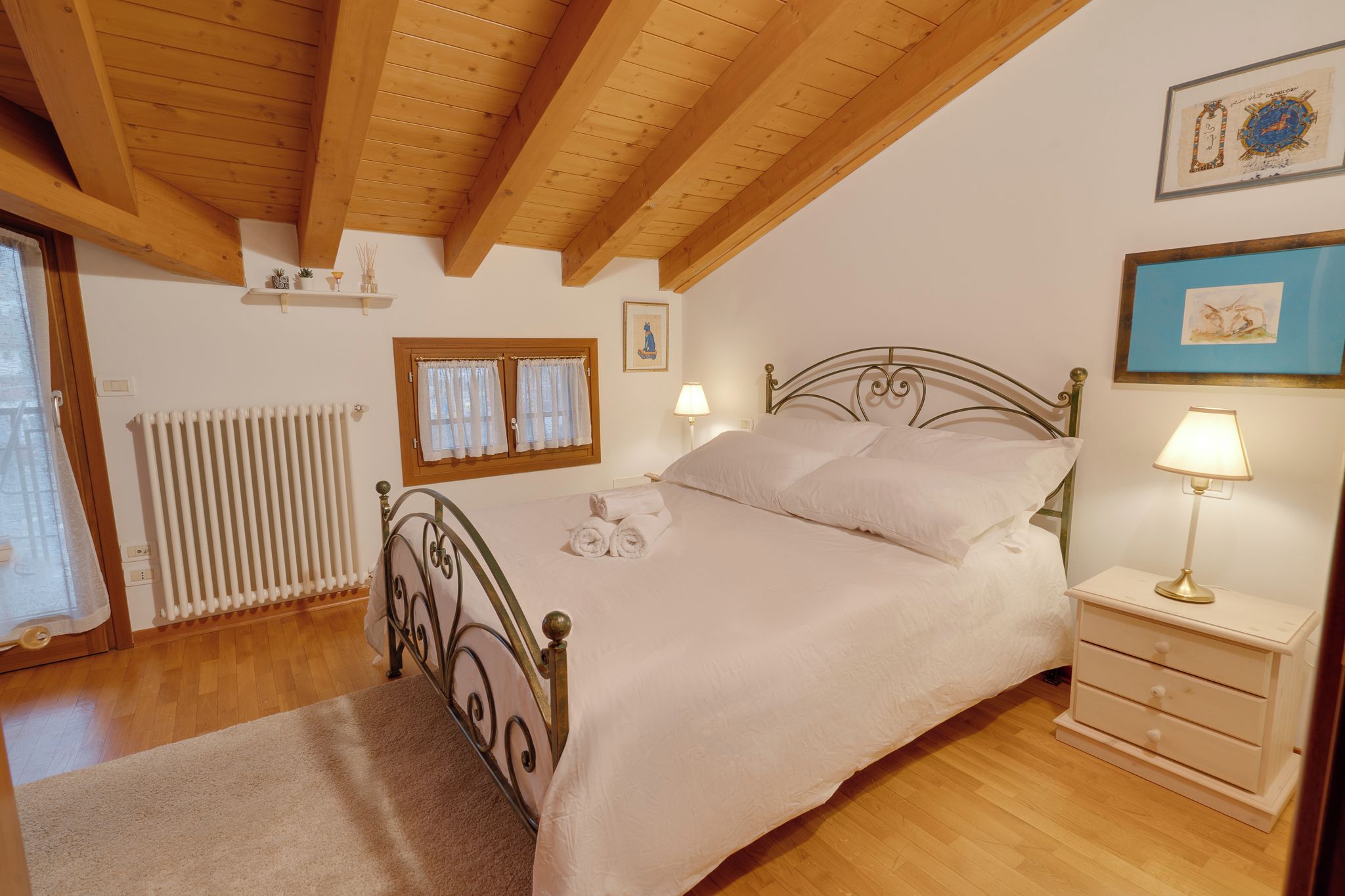Une chambre à coucher comprenant un lit, des tables de chevet, un radiateur, une fenêtre avec des rideaux et des œuvres d’art murales.
