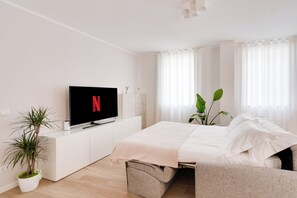 A modern bedroom with a flat-screen TV displaying the Netflix logo, a bed with white bedding, a bedside table, a potted plant, and a window with sheer curtains.